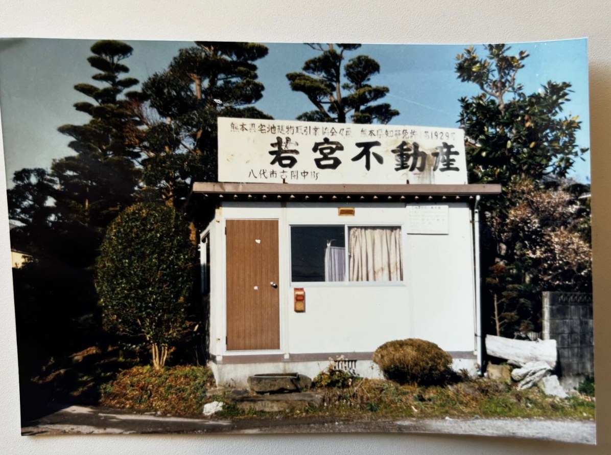 昭和51年撮影・創業当時の若宮不動産の事務所の写真。手書き看板に「熊本県宅地建物取引業協会員 熊本県知事免許（1）第1929号 若宮不動産 八代市古閑中町」とある